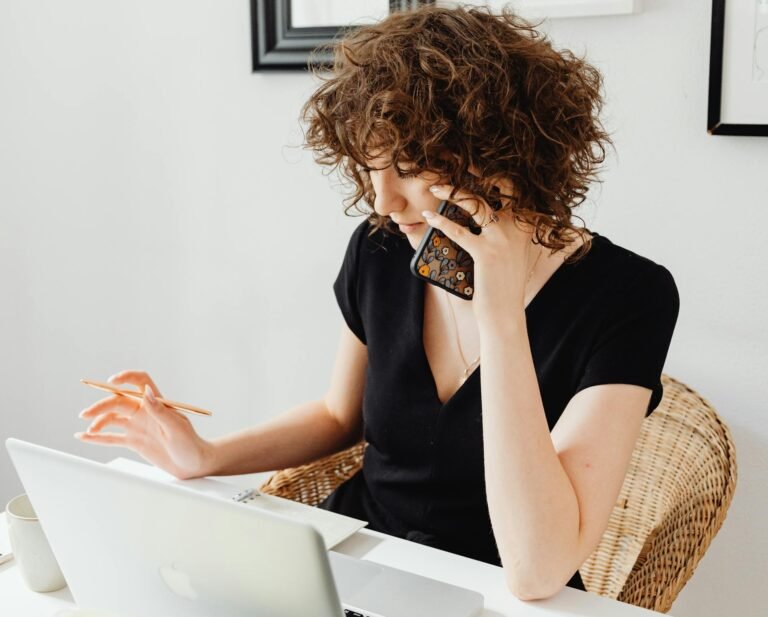 Woman sitting at table, talking on phone while working on laptop in a home office setting.